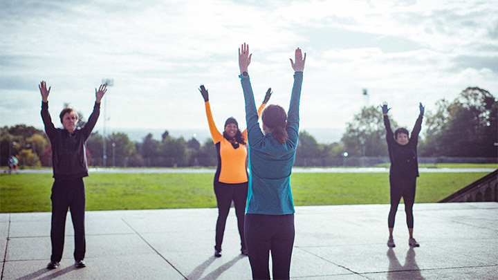 Four people stretching outdoors in a park, raising their hands to the sky.