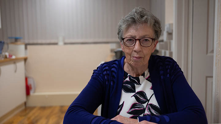 An elderly woman wearing glasses and a blue cardigan over a white shirt sits at a table, looking thoughtfully at the camera.