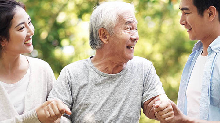 A young couple happily assists an elderly man outdoors, all smiling joyfully.